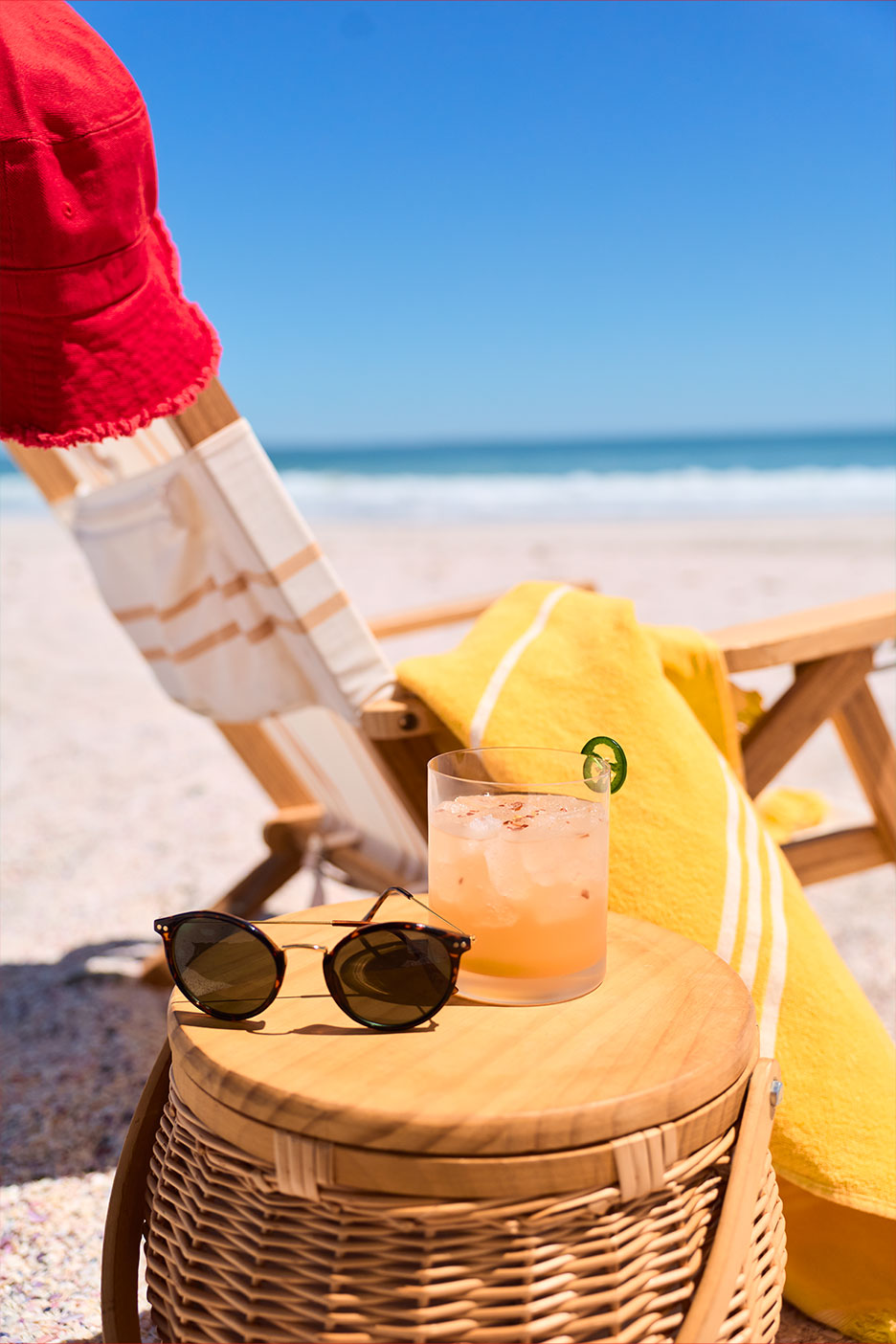 Sonnenbrille und Cocktail auf einem Picknickkorb, mit Strandstuhl und Meer im Hintergrund.