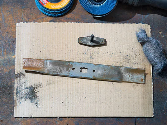 Rostiges Rasenmähermesser auf Karton mit Schleifscheiben und Stahlwolle.