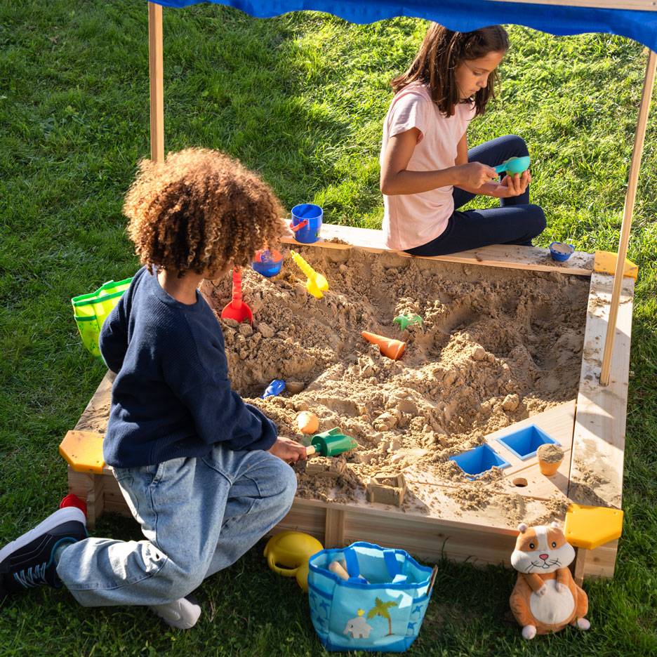 Zwei Kinder spielen in einem Sandkasten mit Dach, umgeben von Sandspielzeug.