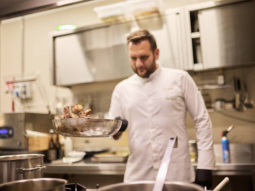 Ein Koch in weißer Kochjacke schwenkt Essen in einer Pfanne.