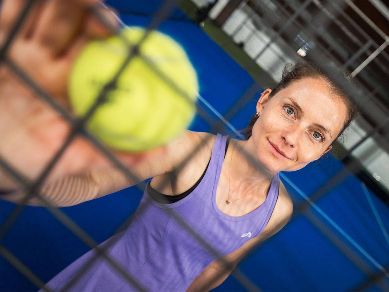 Frau in Sportkleidung hält Padelball durch Netz, blauer Platz im Hintergrund.