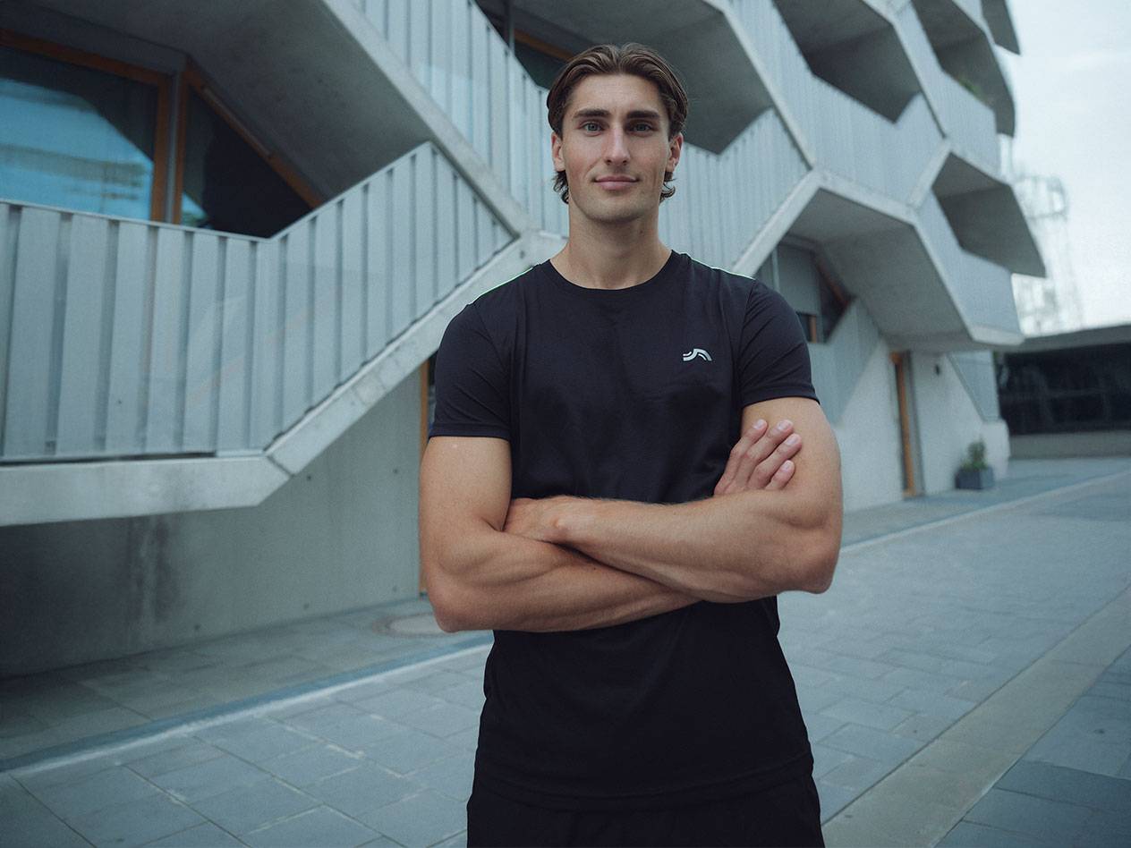 Mann in schwarzem T-Shirt mit Logo und schwarzen Shorts, steht draußen mit verschränkten Armen.