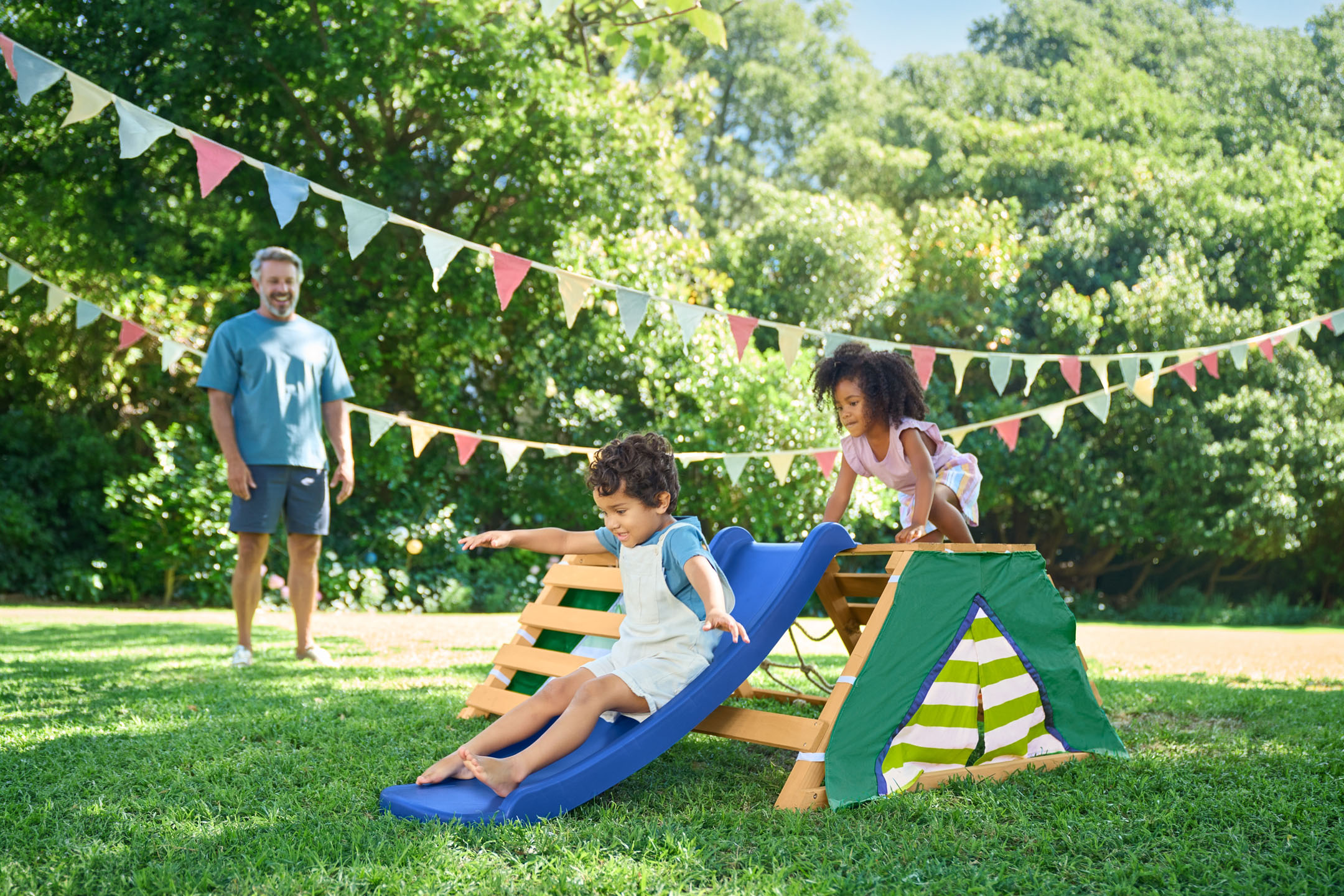 Kinder spielen auf einem Klettergerüst mit Rutsche im Garten, ein Mann im Hintergrund.