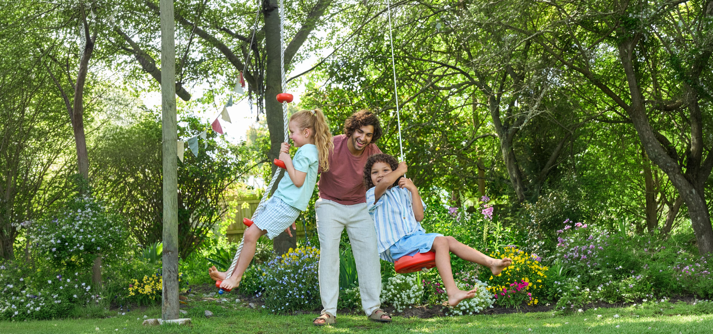 Vater und zwei Kinder spielen auf Schaukeln in einem üppigen grünen Garten.
