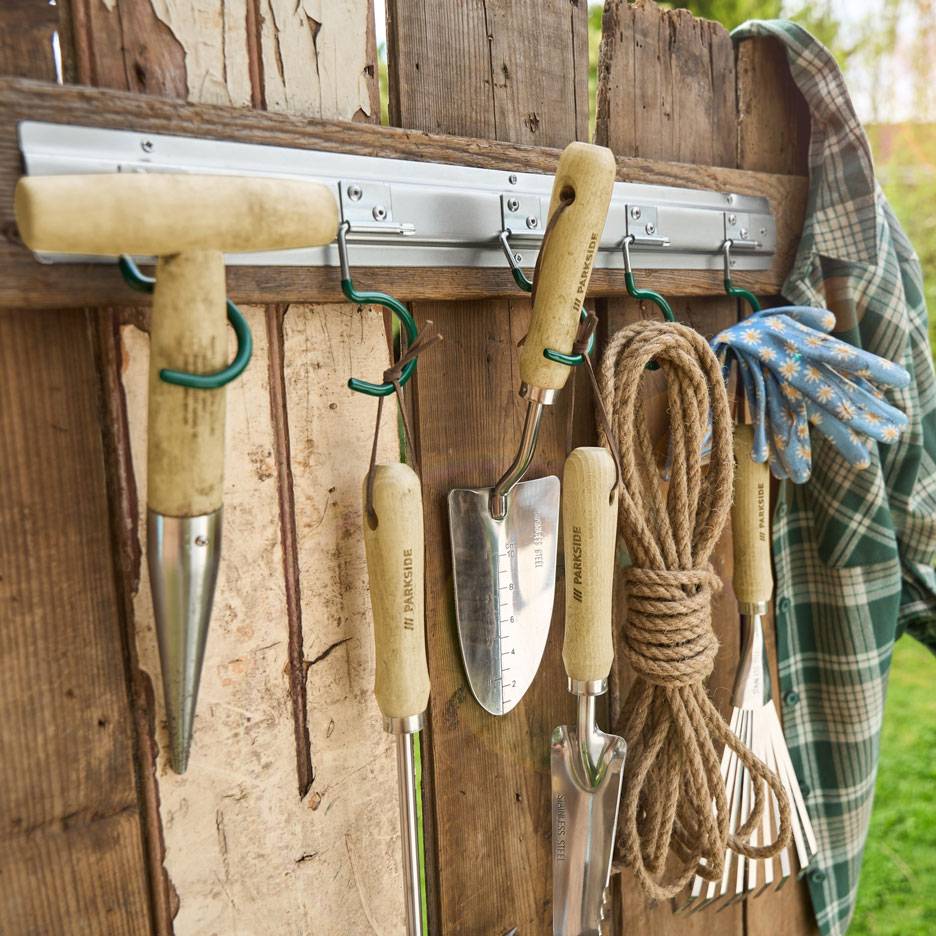 Gartengeräte und Seil hängen an einer Holzwand, neben einem Hemd und Handschuhen.