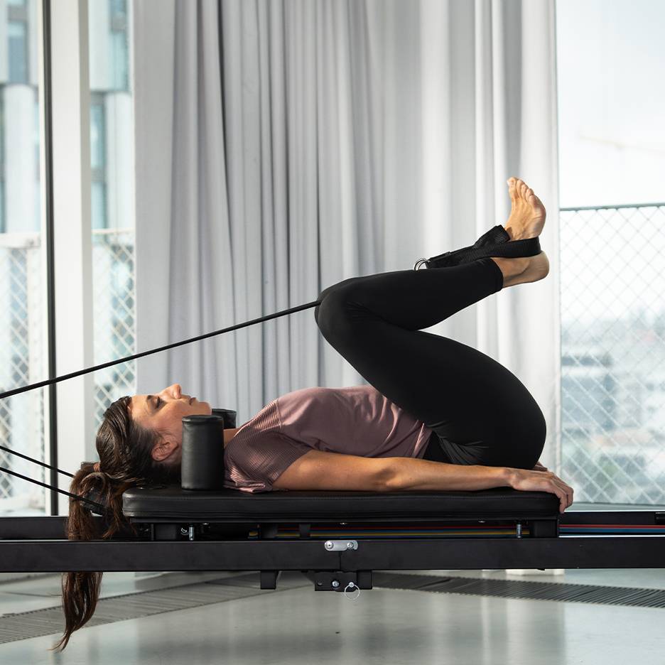 Frau macht Pilates auf einem Reformer mit hochgelegten Beinen.