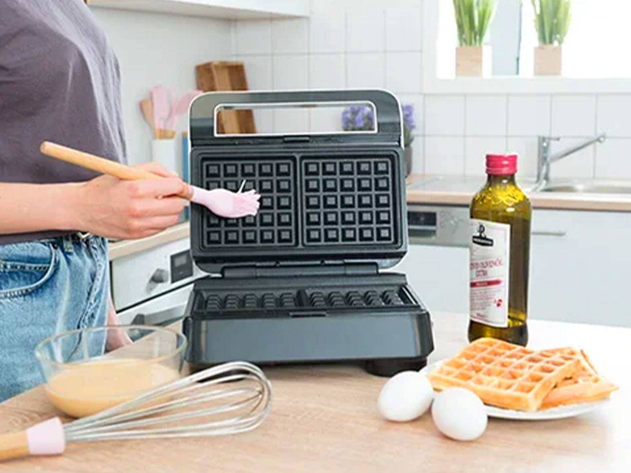 Eine Person bestreicht ein Waffeleisen mit einem Pinsel, mit Zutaten und fertigen Waffeln auf dem Tisch.