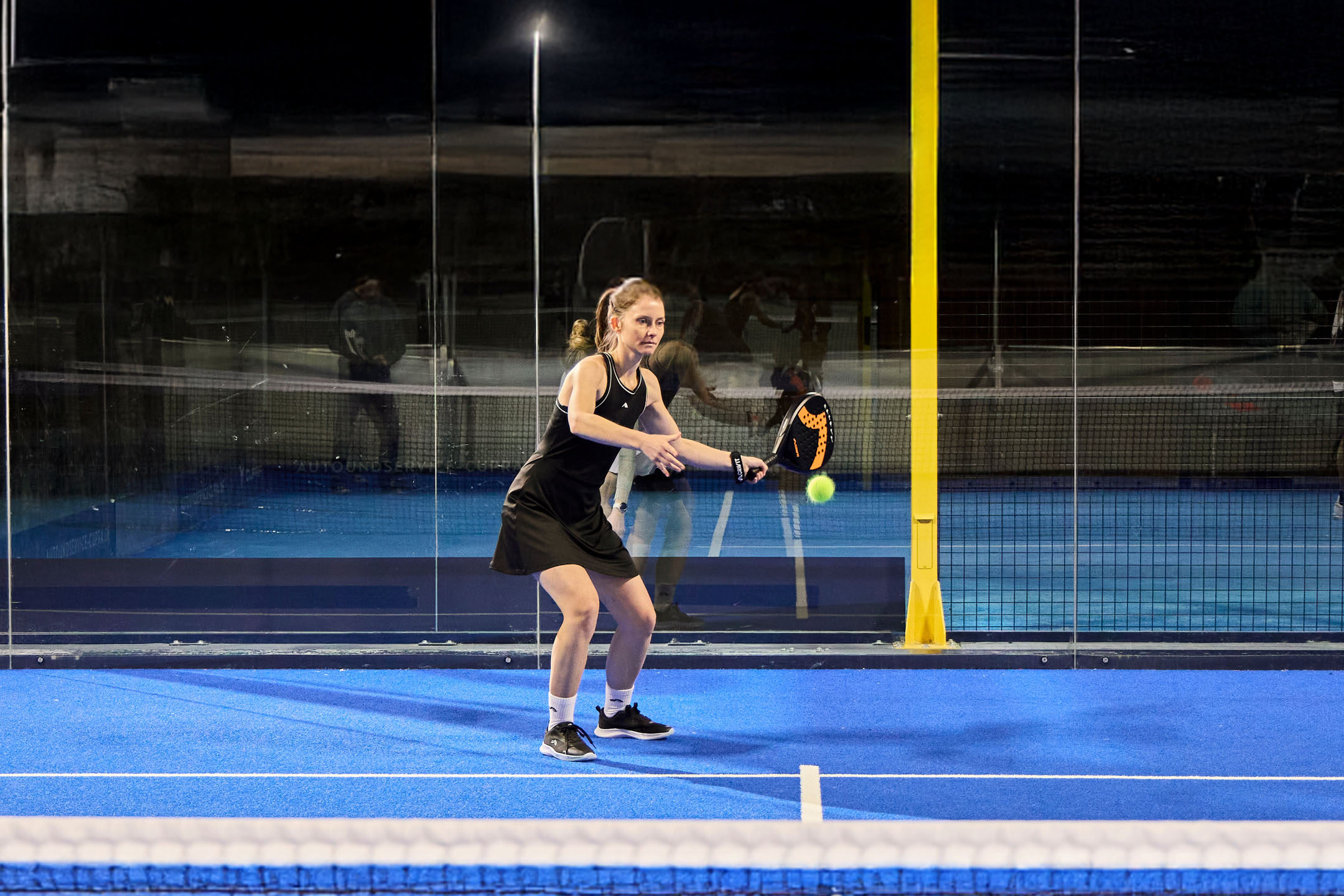 Frau in Sportkleidung spielt Padel auf einem blauen Platz mit Schläger und Ball.