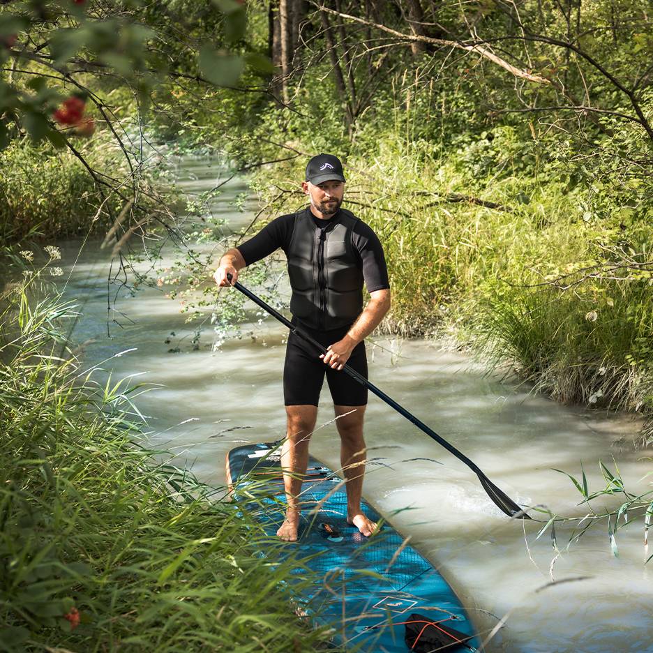Mann in Neoprenanzug und Schwimmweste steht auf einem Paddleboard in einem Bach.