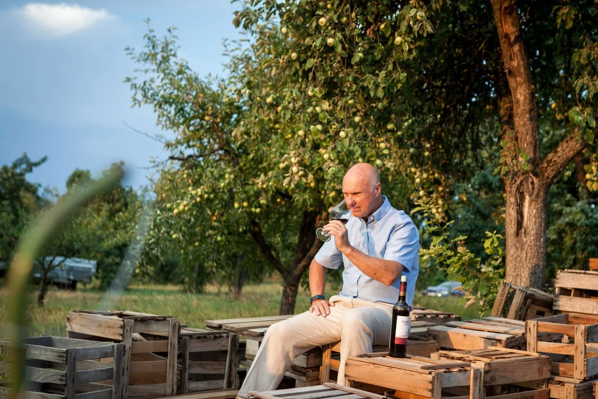 Mann verköstigt Wein inmitten von Holzkisten in einem Obstgarten.