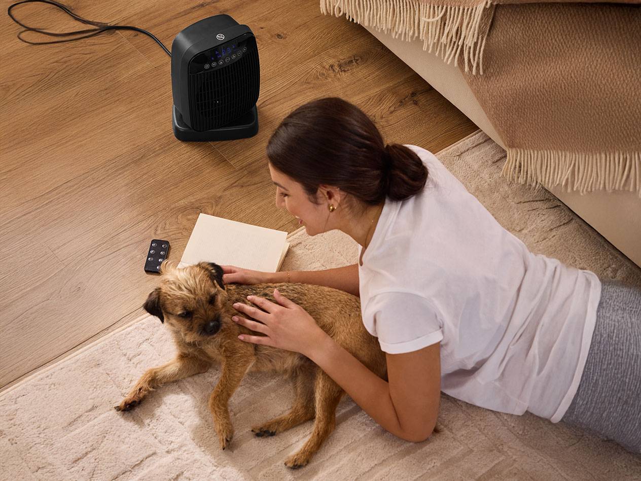 Frau streichelt Hund neben schwarzem Heizlüfter auf dem Boden.