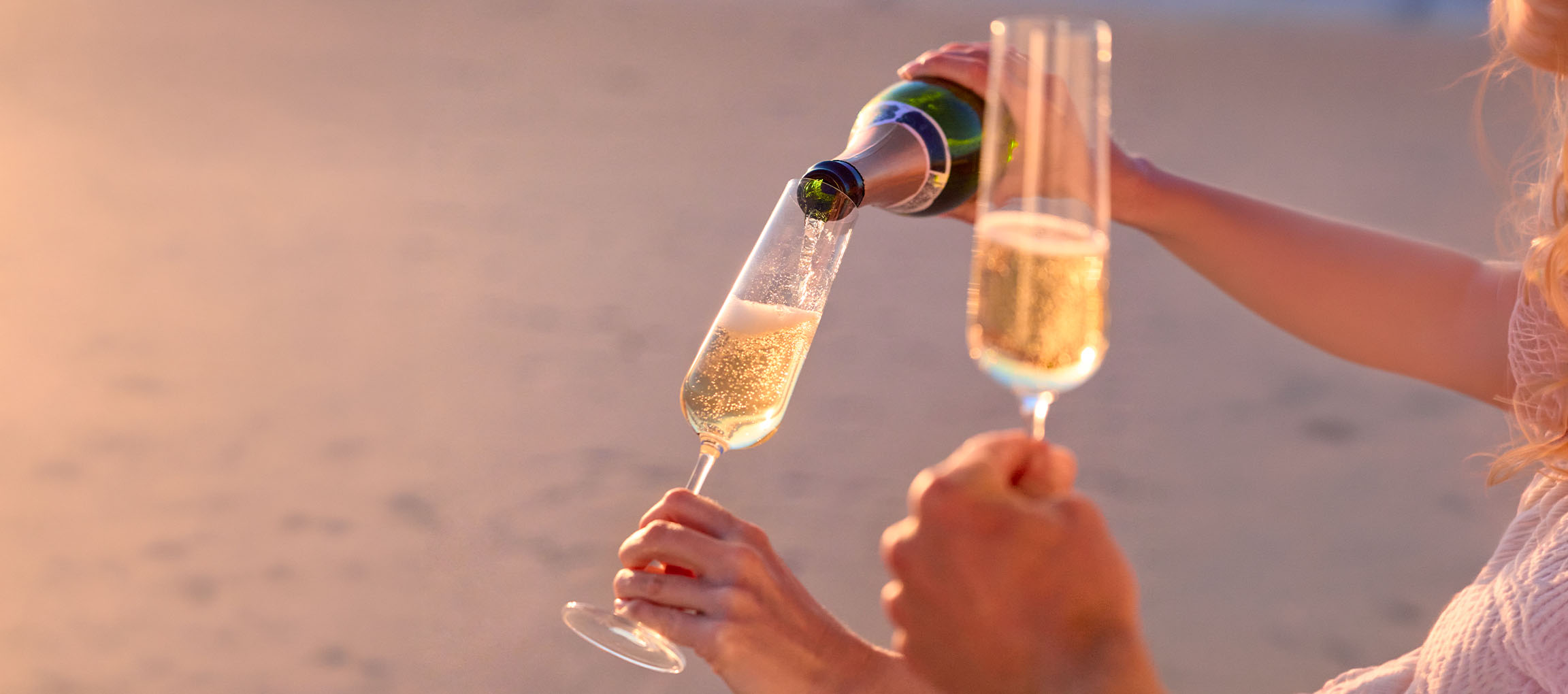 Eine Person gießt Sekt in Gläser am Strand bei Sonnenuntergang.