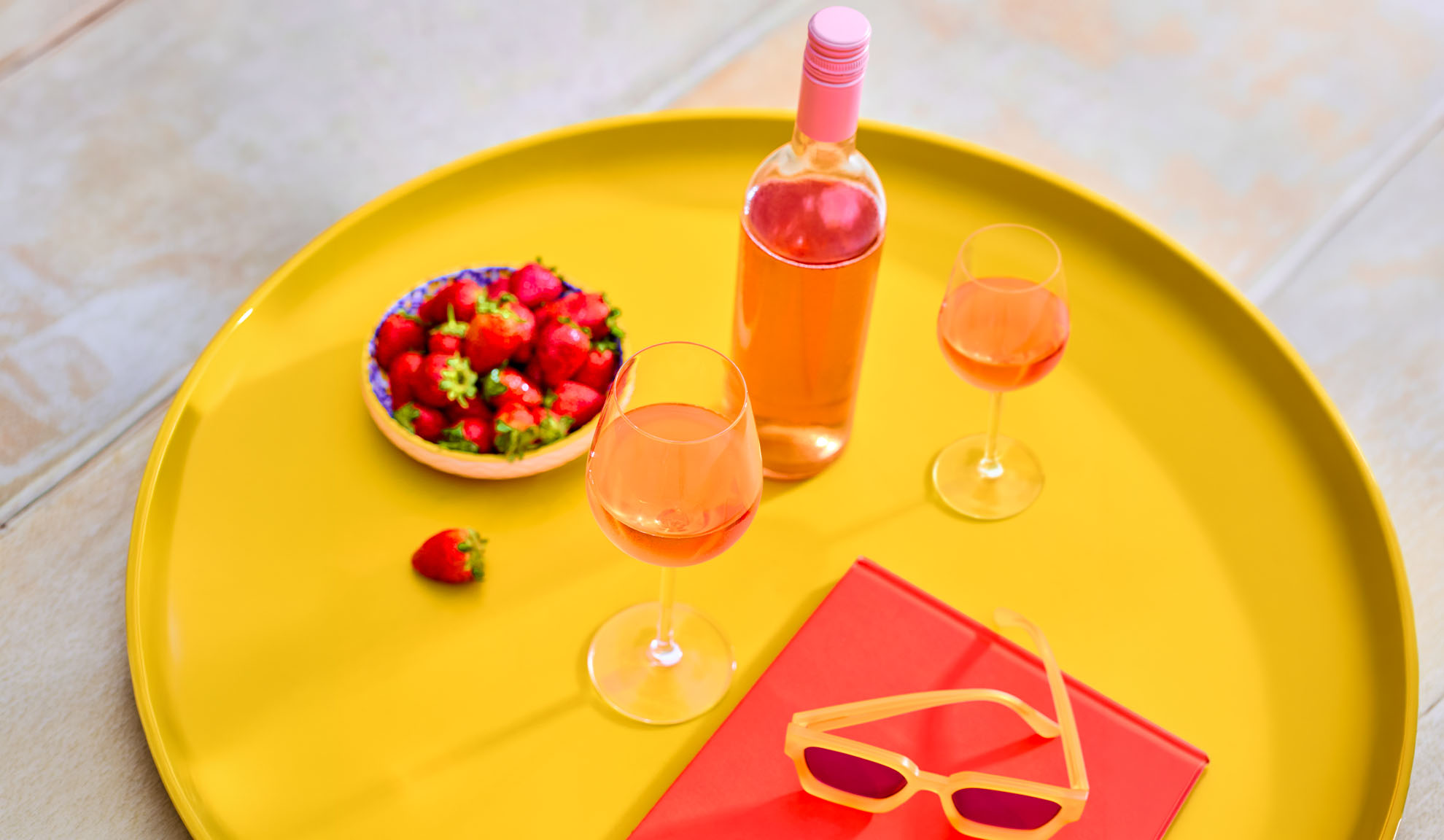 Roséwein, Erdbeeren und Sonnenbrille auf einem gelben Tablett, perfekt für sommerliche Entspannung.