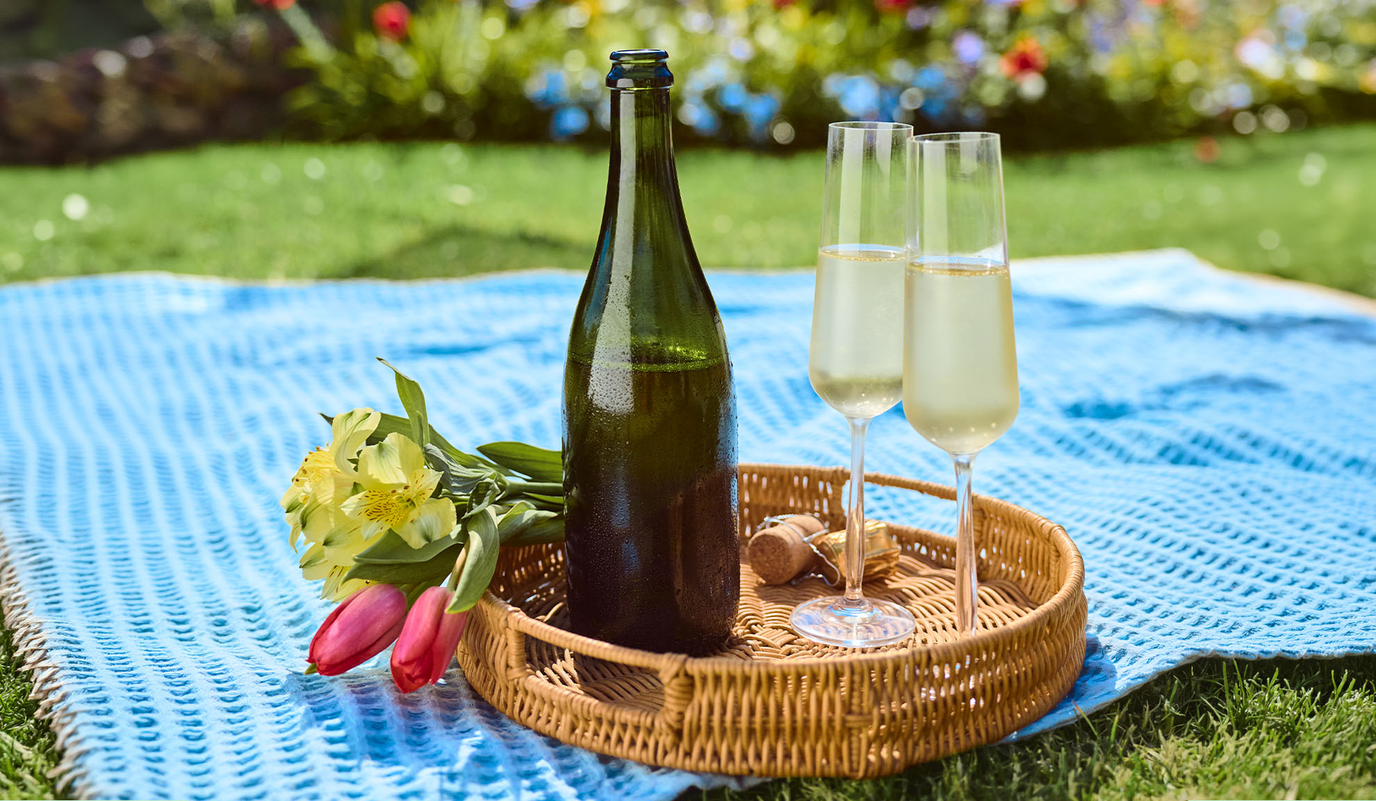 Sektflasche, zwei Gläser und Blumen auf einer blauen Picknickdecke.