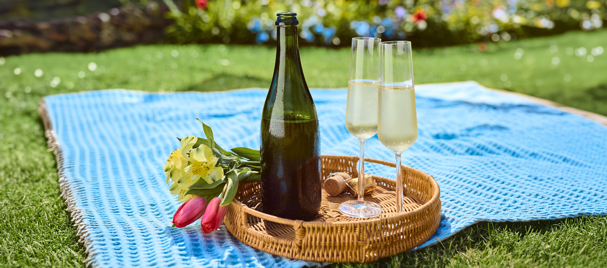 Sektflasche, zwei Gläser und Blumen auf einer Picknickdecke im Garten.