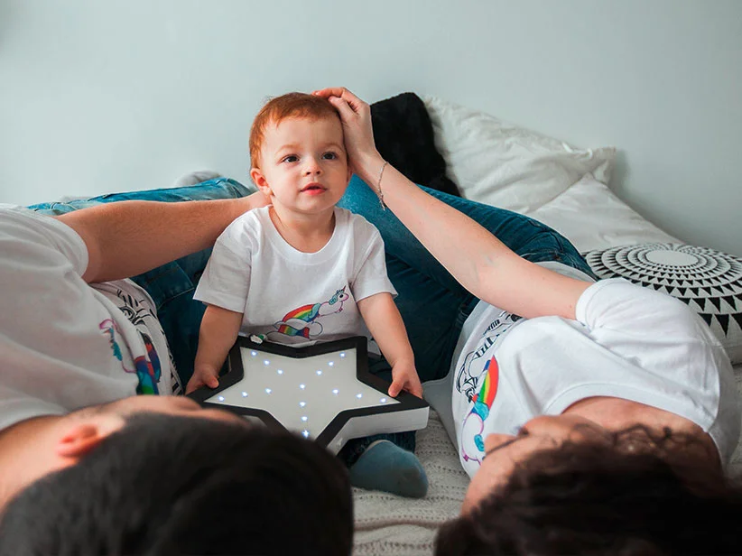 Familie entspannt sich im Bett, Kind trägt Regenbogen-Shirt und Stern-Licht.