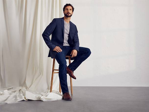 Mann sitzt, trägt dunkelblauen Blazer und Jeans, Lederschuhe.