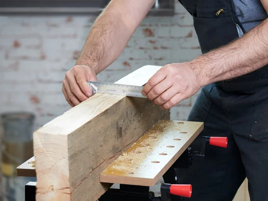 Ein Zimmermann misst ein Holzstück mit einem Metalllineal auf einem Werktisch.