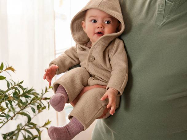 Baby in beigem Kapuzenoverall, gehalten von einem Erwachsenen in grünem T-Shirt.