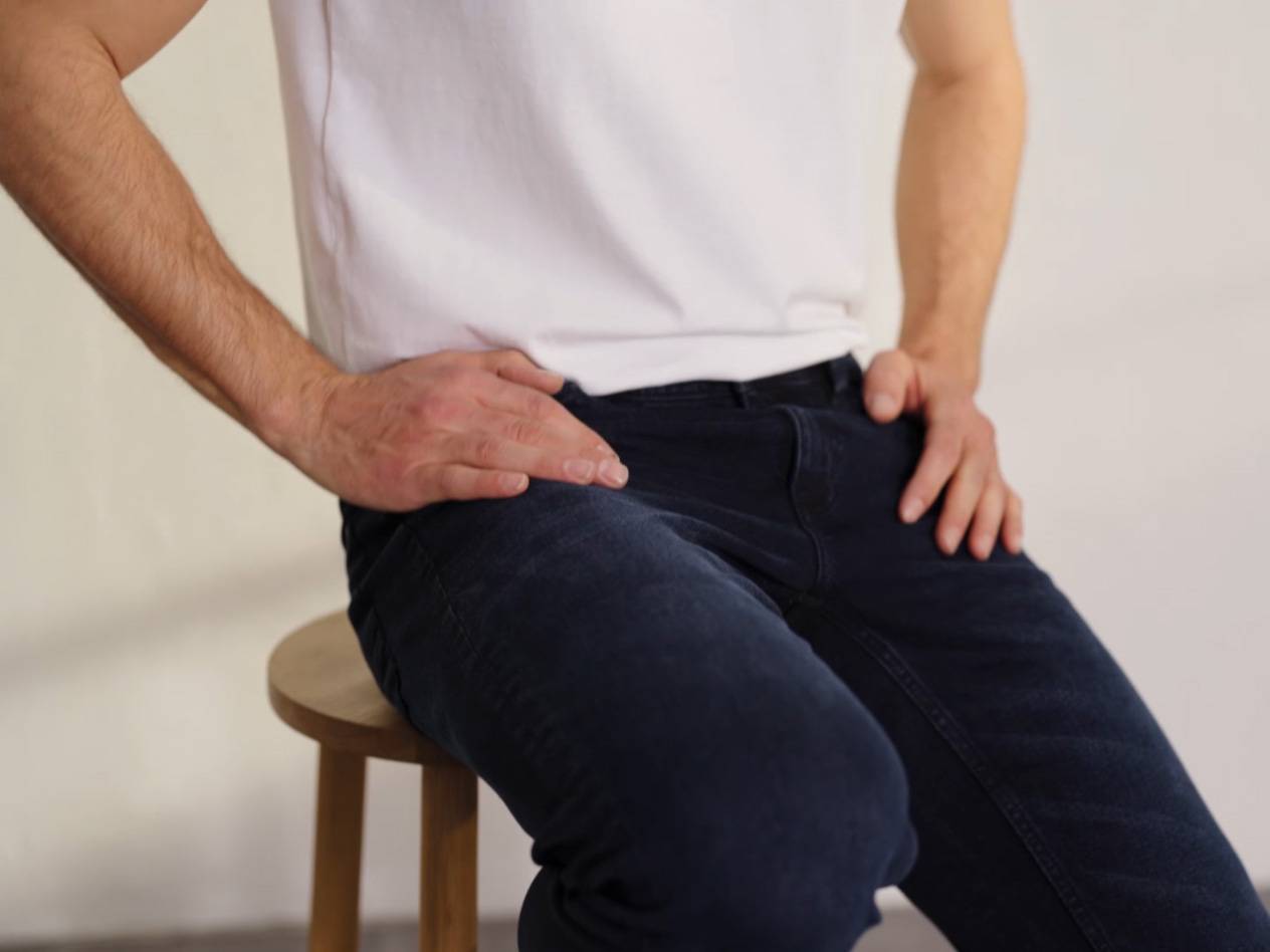 Mann sitzt auf einem Holzhocker, trägt ein weißes T-Shirt und dunkelblaue Jeans.