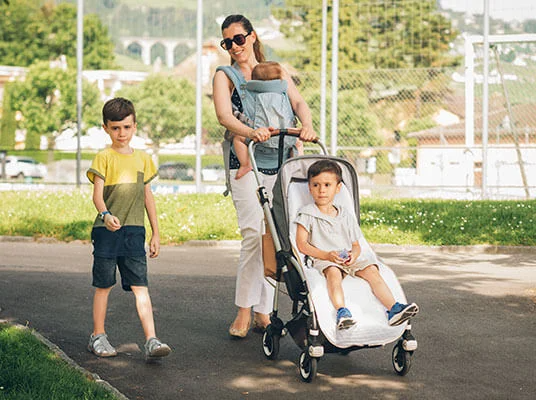 Eine Mutter spaziert mit ihren Kindern im Park, eines im Kinderwagen, eines in der Tragehilfe.