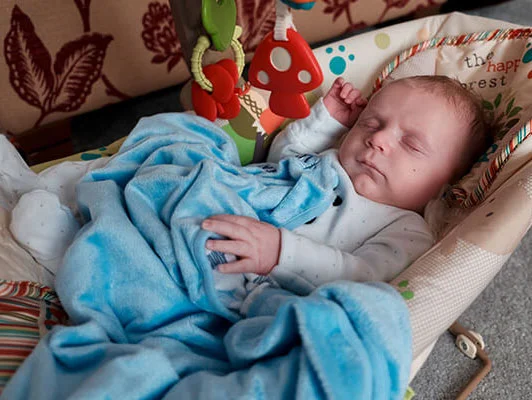 Baby schläft in einem Babywippe, zugedeckt mit einer Decke und Spielzeug.