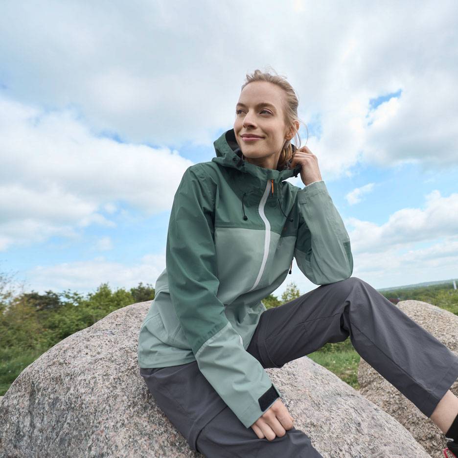 Frau in grüner Outdoor-Jacke und grauer Hose sitzt auf einem Felsen und blickt in die Ferne.