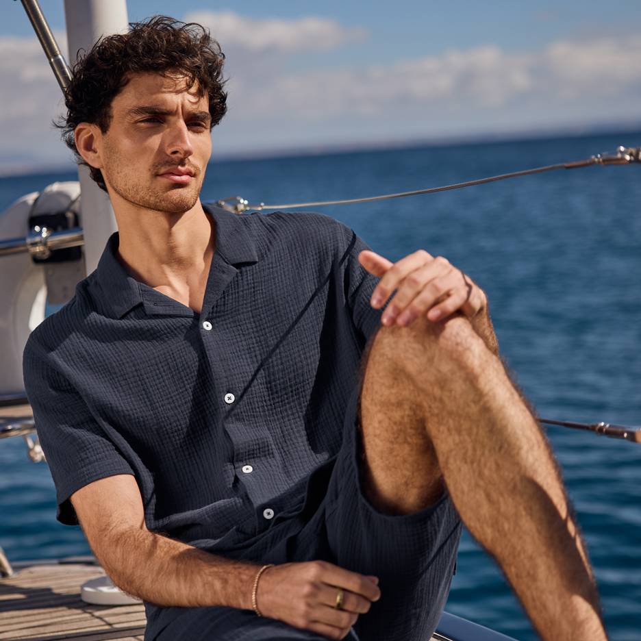 Mann in dunkelblauem Hemd und Shorts auf einer Yacht, blickt aufs Meer.