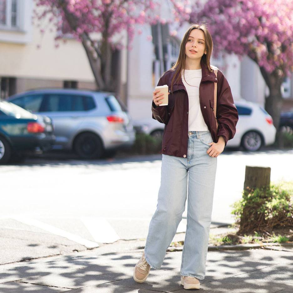 Frau in Jacke und Jeans mit Kaffee auf einer Straße mit blühenden Bäumen.