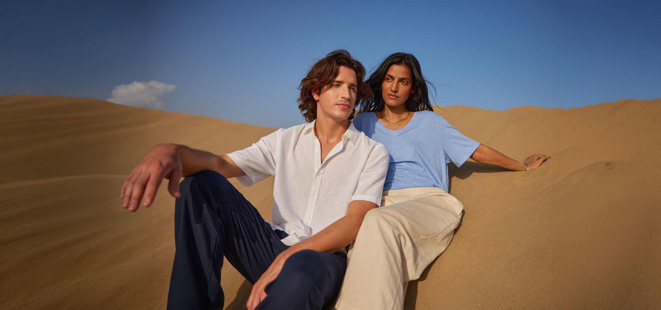 Mann und Frau sitzen auf Sanddünen, tragen Leinenhemden und Hosen.